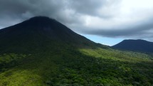 Aerial shot drone flies toward two cloud-covered volcanoes in middle of lush green forest