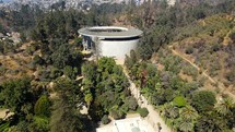 Aerial shot drone flies up and forward toward round music venue and theater hidden in park trees