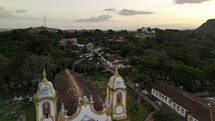 Drone flies backwards over the roof of Igreja de Santo Antonio in Tiradentes, Brazil with the sun setting behind the Serra do Sao Jose