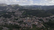 Drone orbits to the left high above the main plaza in Ouro Preto with its rolling hills under cloud cover on a late afternoon