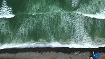 Aerial shot drone flies to right with camera facing down over rocky beach with waves crashing