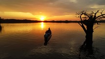 Aerial shot drone slowly rises behind silhouetted tree and canoe boat on black lagoon in middle of Amazon rainforest at sunset
