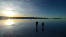 Aerial shot drone flies to right as silhouette couple walk left through water on salt flats at sunrise