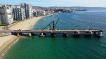 Aerial shot drone flies forward over pier with large antique crane jutting out from sandy beach over green blue waters