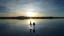 Aerial shot drone flies to right as silhouette couple walk through water on salt flats at sunrise