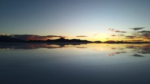 Aerial shot drone ascends over salt flat with reflection in water as sun sets in the distance
