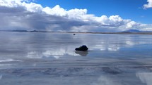 Aerial shot drone flies to left and follows jeep driving on salt flats with reflective water showing a blue sky with fluffy white clouds