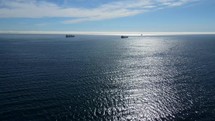 Aerial shot drone hovers over dark blue wavy waters with sun reflection and two ships in the distance on a sunny day with blue skies