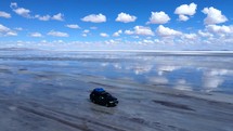 Aerial shot drone orbits to right around jeep driving on salt flats with reflective water showing a blue sky with fluffy white clouds