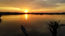 Aerial shot drone hovers above silhouetted tree and canoe boat on black lagoon in middle of Amazon rainforest at sunset