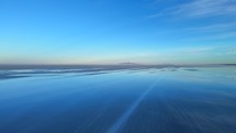 Aerial shot drone flies away from sunrise low over salt flats toward snow capped mountain in the distance
