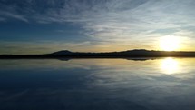 Aerial shot drone flies low and fast over water in salt flats toward mountains with the sunrise behind them