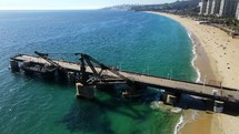 Aerial shot drone flies forward over pier jutting out from sandy beach over green blue waters