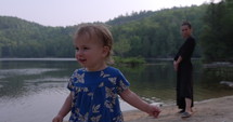Young toddler girl walks along small lake side beach with mother in morning - child exploration