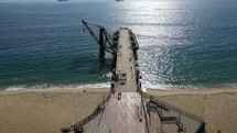 Aerial shot drone flies the length of pier jutting out from sandy beach over green blue waters