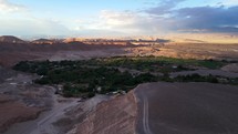 Aerial shot drone flies left over green space in valley below red desert viewpoint at sunset