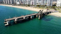 Aerial shot drone orbits to right around pier jutting out from sandy beach over green blue waters
