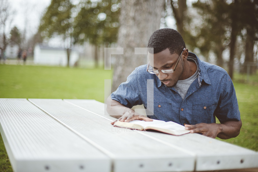 a man reading a Bible sitting at a picnic table in a park 