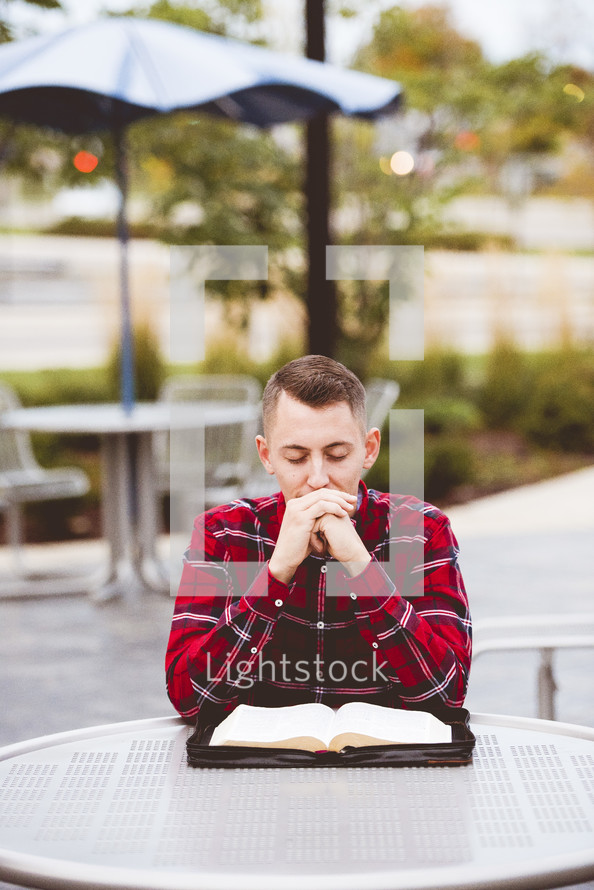 young man praying and reading a Bible sitting at a table outdoors 