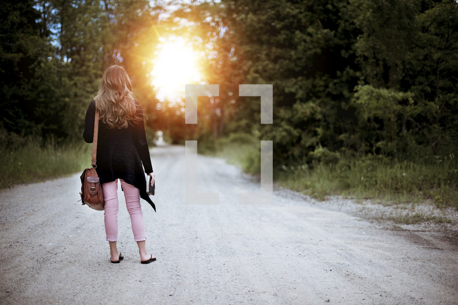 a woman standing in the middle of a dirt road carrying a Bible 