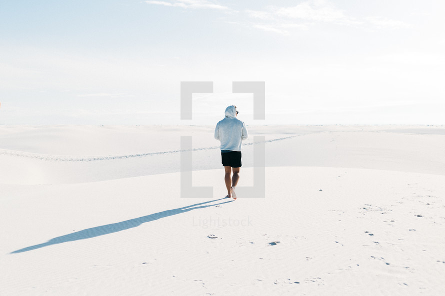 A single man wearing a hoodie walking through a sand filled desert barefoot