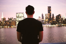 a man looking across a river a city lights at night 