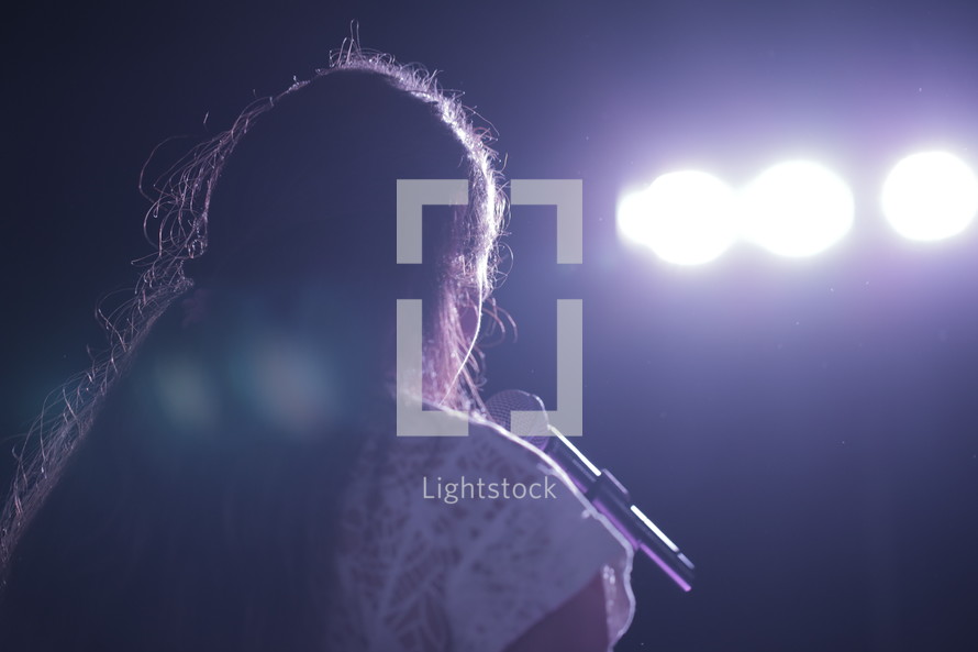 Woman standing at a microphone in front of lights.