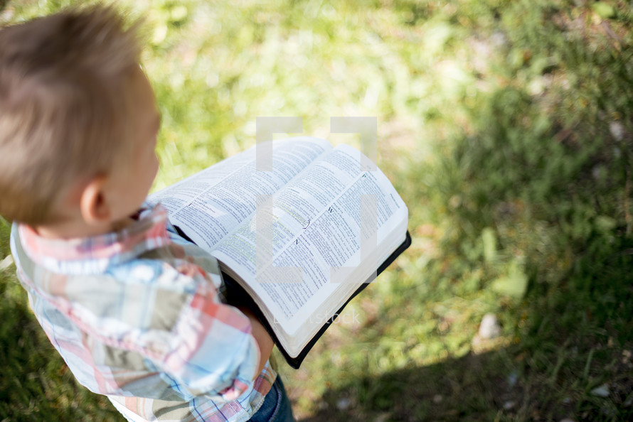 a toddler standing outdoors reading a Bible 