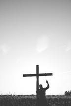 man standing in a field next to a cross with hands raised 