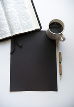 opened Bible, coffee, blank black paper, and pen 
