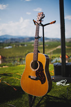 Accoustical guitar on stand in grassy yard with blue sky and mountains on the horizon.