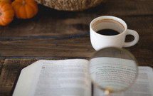 magnifying glass over the pages of a Bible, coffee mug, and pumpkins 