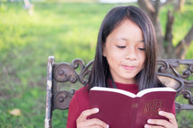 a girl reading a Bible sitting on a park bench 