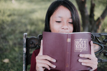 a girl reading a Bible on a park bench 
