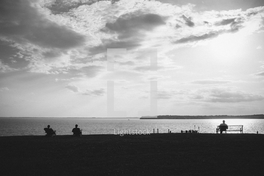 silhouettes of people in chairs sitting at a lake shore 