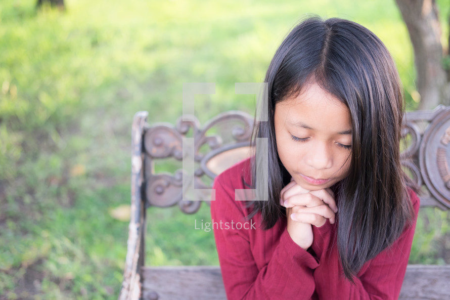 a girl praying sitting on a park bench 