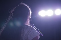 Woman standing at a microphone in front of lights.