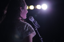 Smiling woman standing at a microphone in front of lights.
