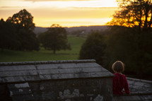 child sitting on a stone wall looking out at a green pasture 