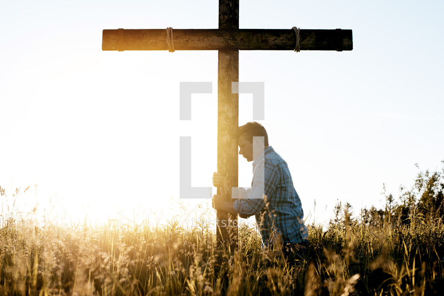 a man standing in a field next to a cross with head bowed 