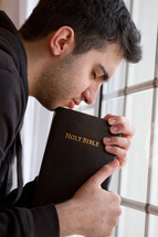 man praying by a window holding a Bible