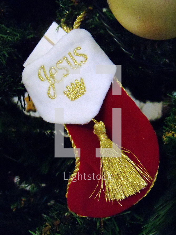 A red and white Christmas stocking with Jesus embroidered on the stocking in gold embroidery lettering waits for Jesus on a Christmas Tree surrounded by Christmas tree ornaments. 