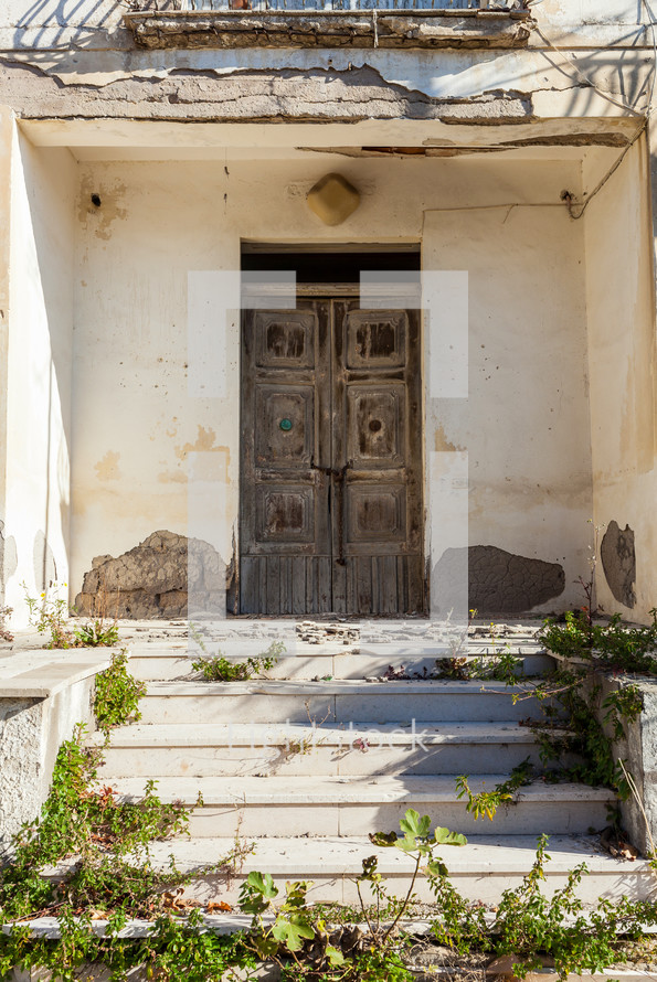 Old wooden door on the facade of an abandoned house with plants growing on the steps