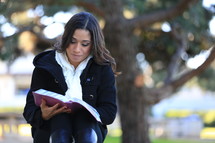 Woman sitting in front of large tree reading Bible.