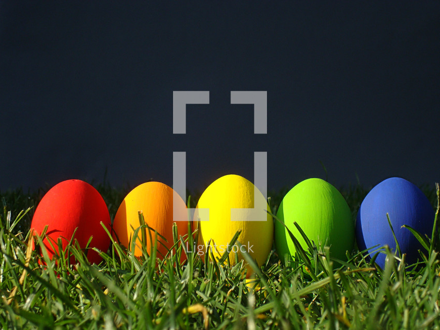 multicolored eggs in the grass in front of a dark gray background. 
