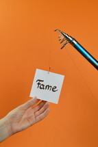 a hand grabbing for a piece of paper with the word FAME on it hanging from a fishing line
