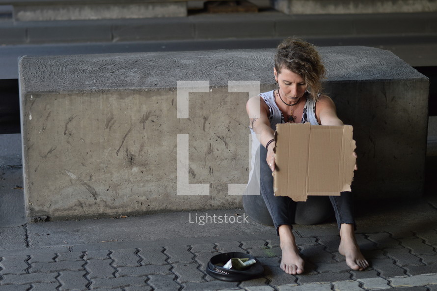 a homeless woman holding a blank sign under a bridge 