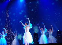 A group of ballet dancers performing the nutcracker ballet at Christmas time in front of a Christmas tree and blue backdrop with snow flakes. 