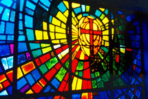 An ornate and colorful stained glass window displaying the Cross of Christ surrounded by red, orange, yellow, green, purple and blue colors in a stained glass window in a church in Louisiana. 