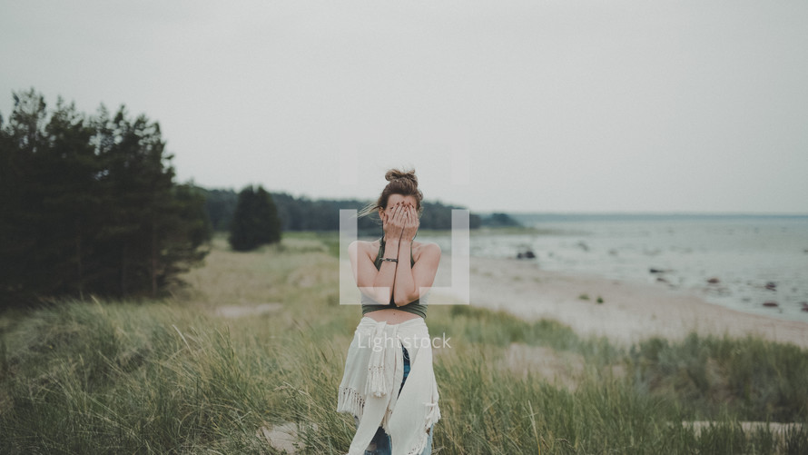 a woman standing on a shore covering her face with her hands 